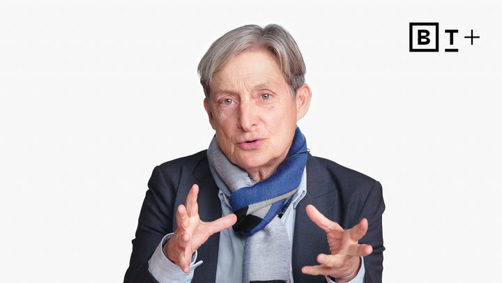 An older person with short gray hair, wearing a suit jacket and scarf, gestures with both hands. A "B T +" logo is visible in the top right corner.