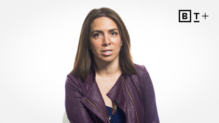 A woman with straight brown hair wearing a purple jacket sits facing the camera against a plain white background with a "BT+" logo in the top right corner.