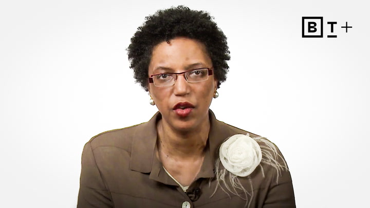 A woman with short curly hair and glasses, wearing a brown suit jacket with a large white flower brooch, speaks in front of a plain white background with a "B T +" logo.