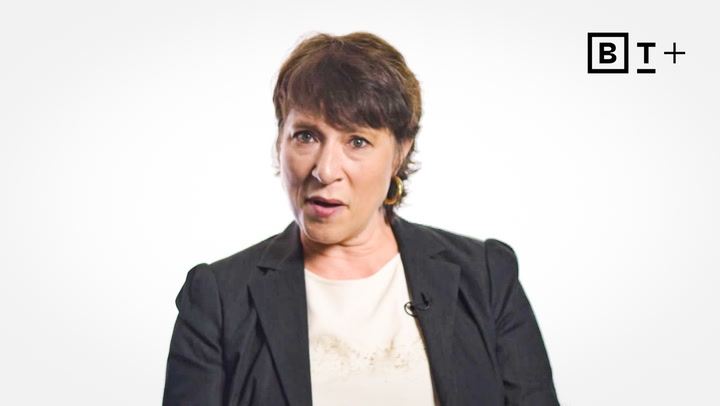 A woman with short dark hair, wearing a black blazer and light top, sits in front of a white background with a "B T +" logo in the top right corner.