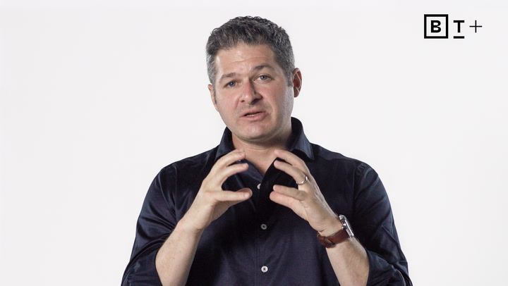 A man with short hair wearing a dark shirt gestures with his hands while speaking against a plain white background. The letters "BT+" are visible in the top right corner.