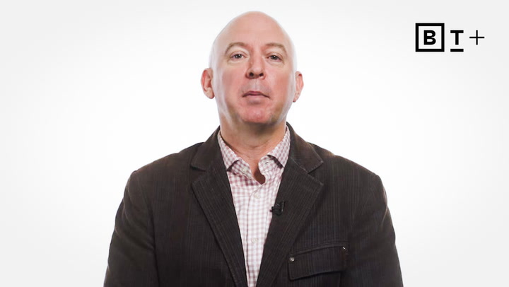 A bald man in a checked shirt and dark blazer sits in front of a plain white background with a "BT+" logo in the top right corner.
