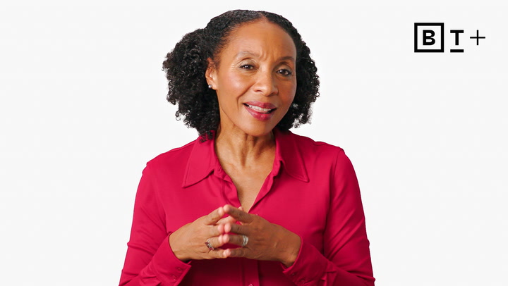 A woman with curly hair wearing a red blouse stands in front of a white background with the letters "BT+" in the top right corner.