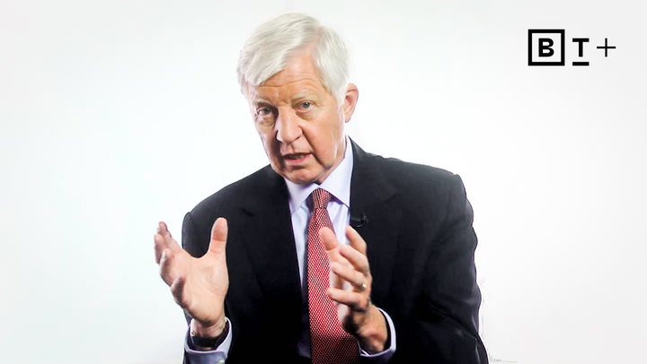An older man in a suit and red tie speaks while gesturing with his hands. A "BT+" logo is visible in the top right corner against a white background.