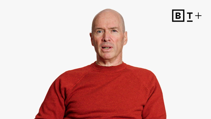 A middle-aged man in a red sweater sits against a plain white background with a "B T +" logo in the top right corner.
