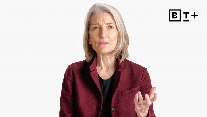A woman with gray hair wearing a burgundy blazer gestures with one hand against a plain white background with a "BT+" logo in the top right corner.