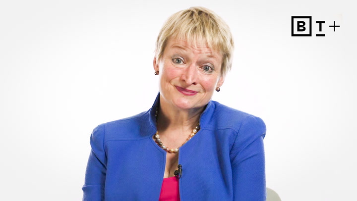 A woman with short blonde hair, wearing a blue blazer and pink top, sits in front of a white background with a "B T +" logo in the top right corner.
