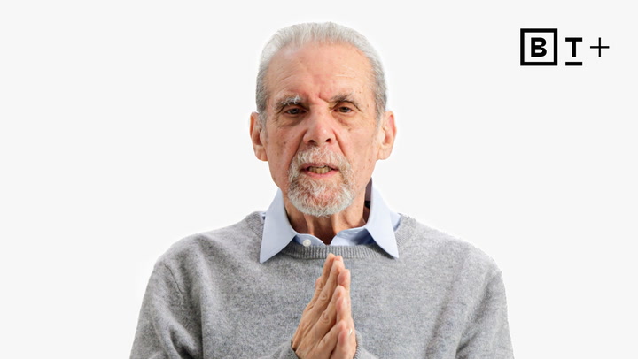 An older man with gray hair and a beard wears a gray sweater over a collared shirt, standing against a white background with a "B T +" logo in the top right corner.
