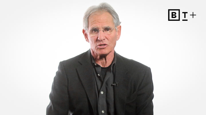 A man with gray hair wearing glasses and a dark suit jacket sits in front of a white background with a "B T +" logo in the top right corner.
