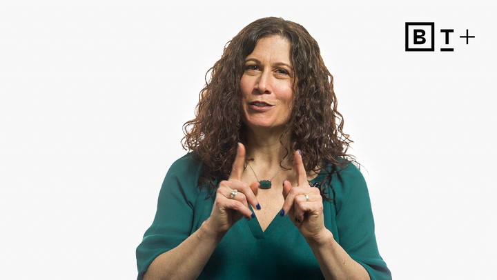A woman with curly brown hair wearing a green blouse gestures with both index fingers raised. A "B T +" logo appears in the top right corner on a white background.