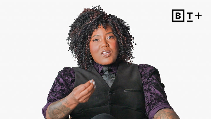 A person with curly hair wearing a purple patterned shirt, black vest, and tie sits against a white background, gesturing with one hand. The letters "BT+" are in the top right corner.
