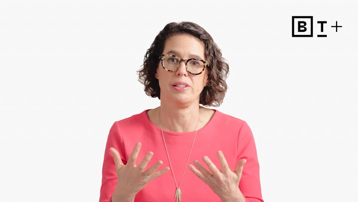 A woman with glasses and curly hair, wearing a pink top, gestures with her hands while speaking in front of a plain white background. A "B T +" logo appears in the top right corner.