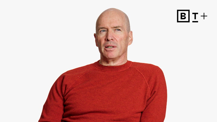 A man in a red sweater sits against a plain white background with a “BT+” logo in the top right corner.