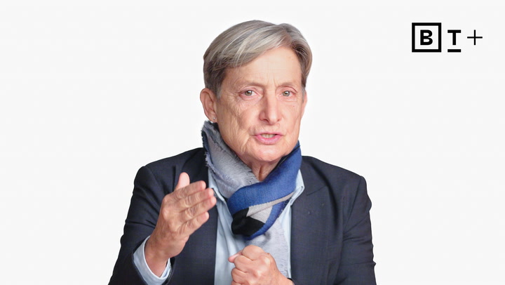 An older person with short gray hair, wearing a blazer and scarf, gestures with one hand. A "BT+" logo appears in the top right corner against a white background.