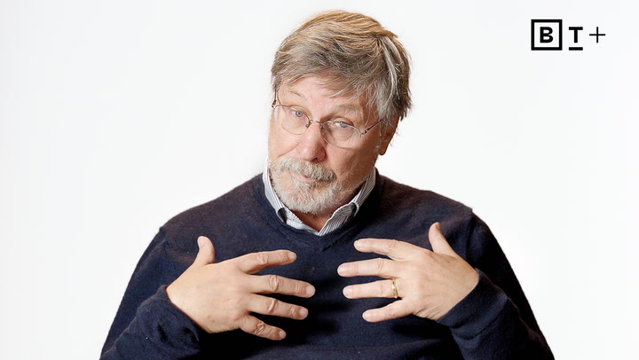 An older man with glasses and a beard, wearing a dark sweater, gestures with both hands on his chest against a plain white background with a "BT+" logo in the corner.