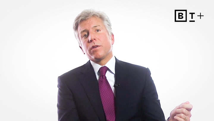 A man in a dark suit and maroon tie speaks while seated against a white background with a "B T +" logo in the top right corner.