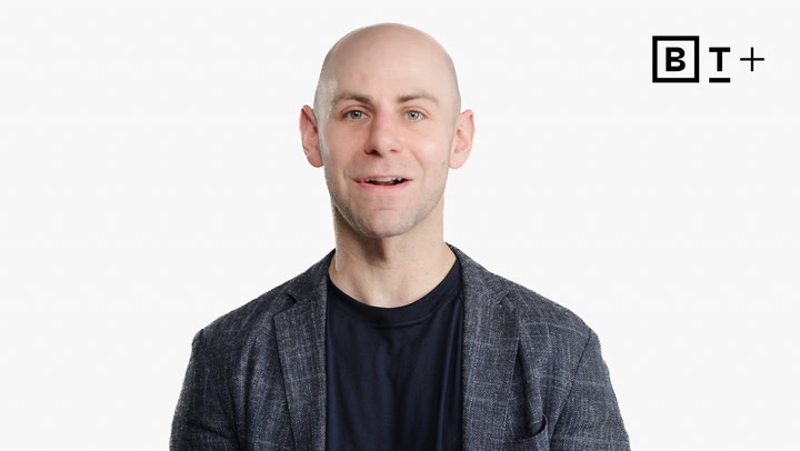 A bald man in a dark shirt and checked blazer stands in front of a white background with a "B T+" logo in the top right corner.