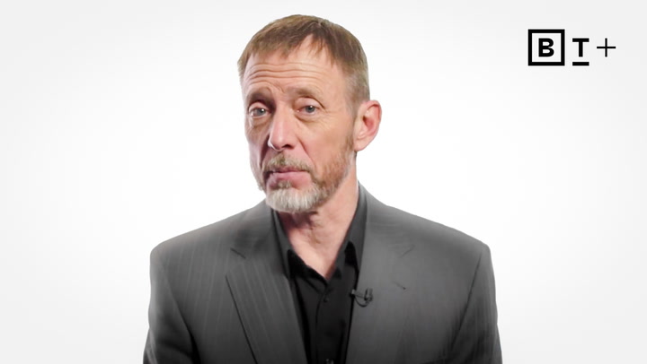 A man with short hair and a beard, wearing a gray suit and black shirt, stands against a plain white background with a "BT+" logo in the top right corner.