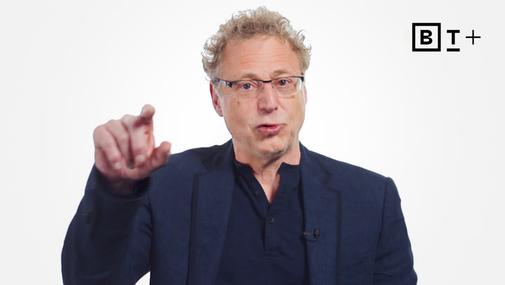 A man with curly blond hair and glasses, wearing a navy blazer and dark shirt, gestures with his right hand against a plain white background with a "BT+" logo in the top right corner.