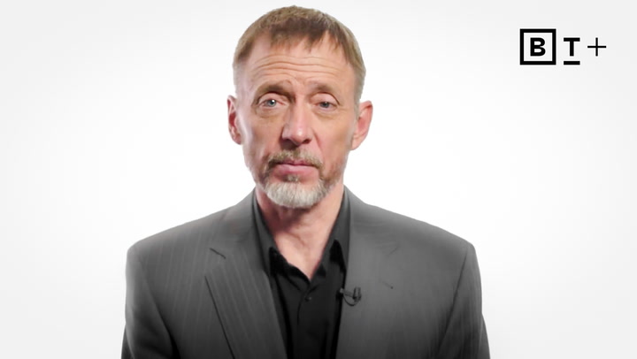 A middle-aged man in a gray suit and black shirt stands against a plain white background with a "BT+" logo in the top right corner.