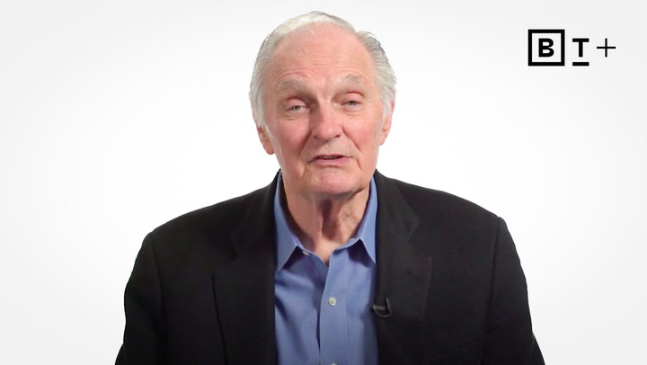 An older man in a blue shirt and black blazer speaks in front of a white background with a "B T +" logo in the top right corner.