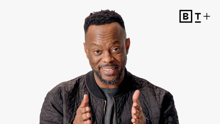 A man with a short beard and black jacket gestures with his hands, standing in front of a plain white background with a "B T +" logo in the corner.