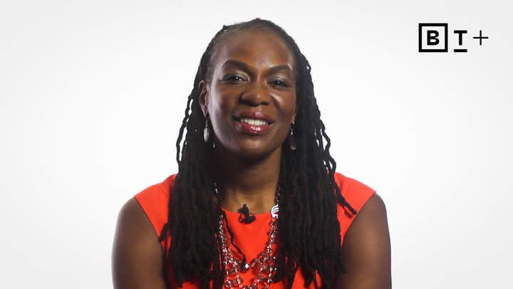 A woman with long braided hair, wearing a bright orange sleeveless top and a necklace, is smiling at the camera against a plain white background with a "B T +" logo in the top right corner.