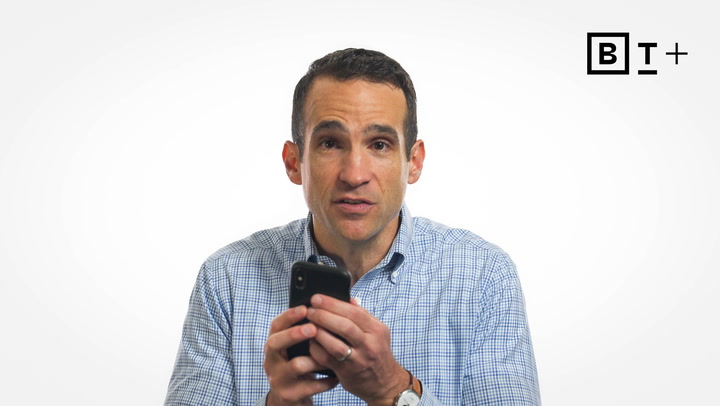 A man in a checkered shirt holds a smartphone and looks at the camera, with a "B T +" logo in the top right corner on a plain white background.