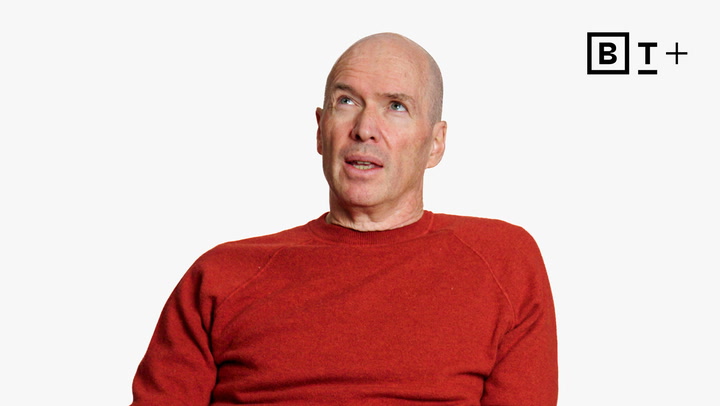 A bald man in a red sweater looks upward against a plain white background, with the "B T +" logo in the top right corner.