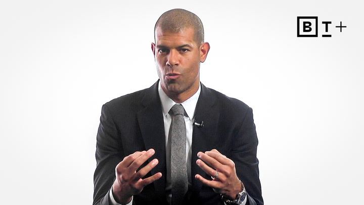 A man in a suit gestures with his hands while speaking, seated against a plain white background with a "B T +" logo in the top right corner.