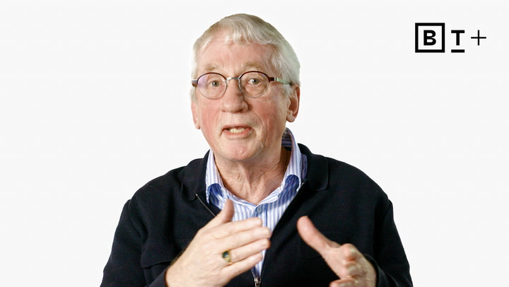 An older man with glasses and white hair gestures while speaking in front of a plain white background with a "B T +" logo in the top right corner.