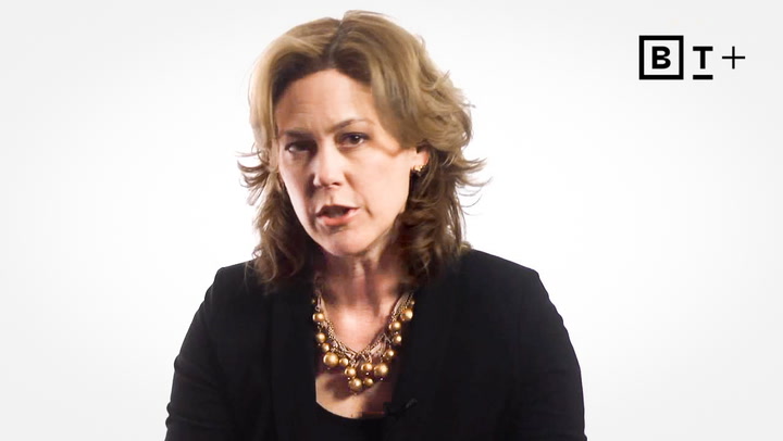 A woman with shoulder-length brown hair wearing a black top and a gold statement necklace sits in front of a plain white background with a "BT+" logo in the upper right corner.