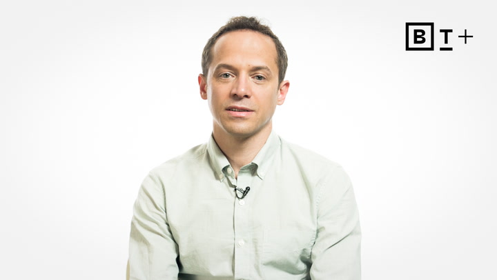 A man in a light-colored shirt sits facing the camera against a plain white background, with a small microphone clipped to his shirt. A "B T +" logo appears in the top right corner.