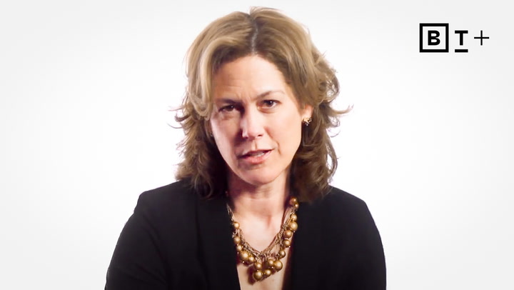 A woman with shoulder-length wavy hair wearing a black blazer and a gold necklace looks at the camera; a "B T +" logo is in the top right corner.