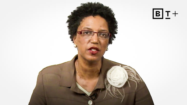 A woman with short curly hair, glasses, and a brown jacket with a white flower pin speaks in front of a plain white background, with the BT+ logo in the top right corner.