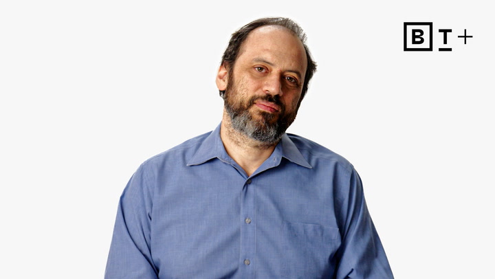 A man with a beard and receding hairline, wearing a blue collared shirt, is facing the camera against a plain white background with a "B T +" logo in the top right corner.