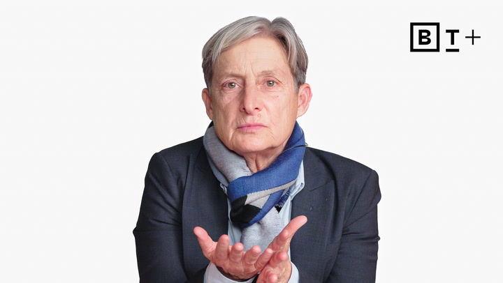 An older person with short gray hair, wearing a dark blazer and scarf, holds their hands out with palms up against a plain white background.