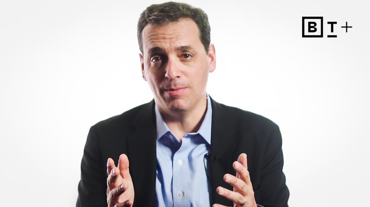 Man in a suit jacket and blue shirt gestures with both hands, sitting in front of a plain white background with a "BT+" logo in the top right corner.