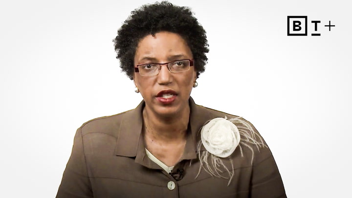 A woman with short curly hair and glasses wears a brown jacket with a large white flower accessory, speaking in front of a plain white background with a "B T +" logo.