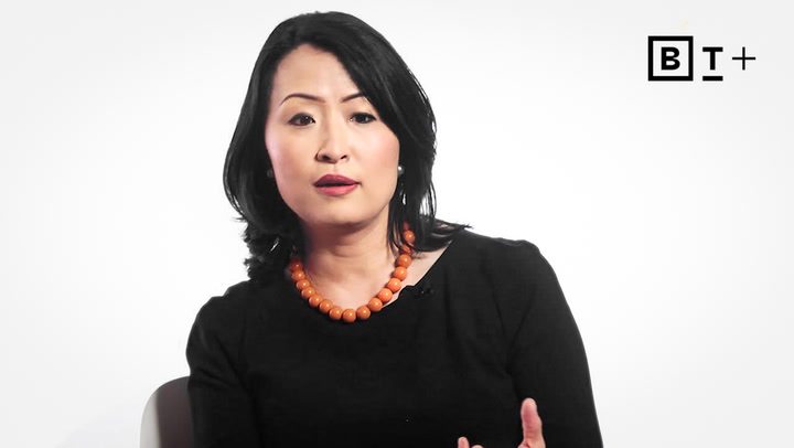 A woman with long black hair, wearing a black top and an orange beaded necklace, sits and speaks against a plain white background with a "B T +" logo in the top right corner.