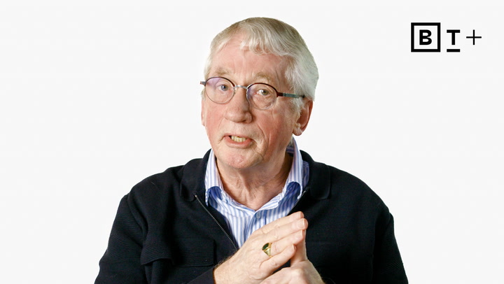 An older man with glasses and white hair speaks, wearing a dark jacket over a striped shirt, with a BT+ logo in the top right corner.