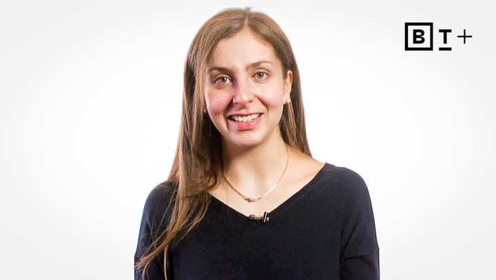 A woman with long brown hair, wearing a black top and a necklace, stands against a plain white background with a "BT+" logo in the top right corner.