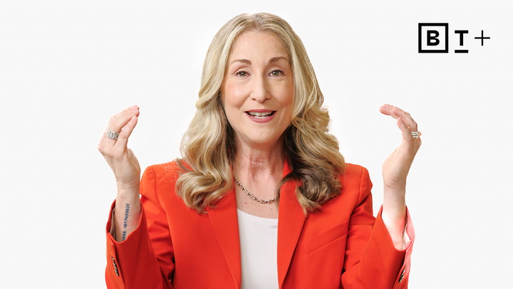 A woman with long blonde hair in an orange blazer gestures with both hands, smiling in front of a plain white background with a "B T +" logo in the top right corner.