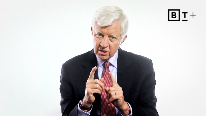 An older man in a suit and red tie speaks, gesturing with both hands. The background is plain white, with a “B T +” logo in the top right corner.