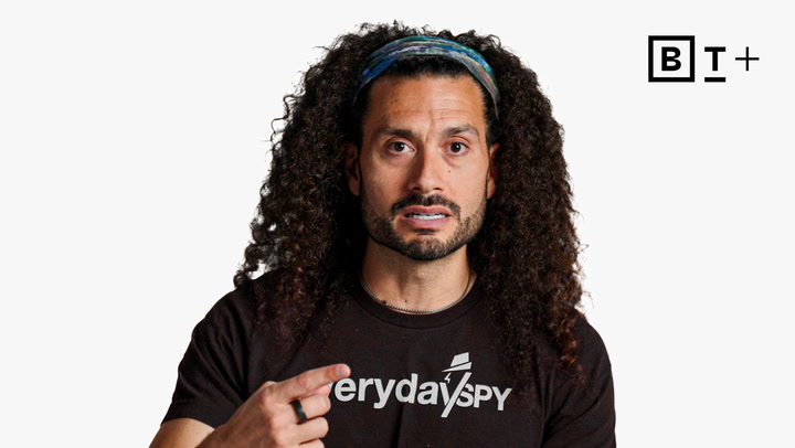 A person with long curly hair and a headband speaks while pointing to themselves, wearing a black "Everyday Spy" shirt. The background is white with a "B T +" logo in the top right corner.