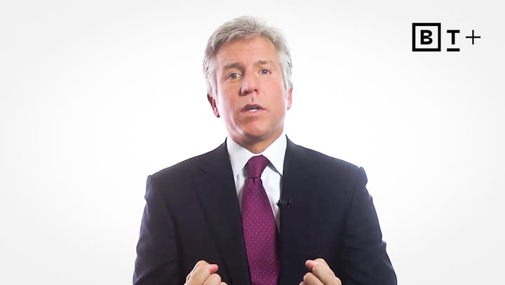 A man in a dark suit and red tie speaks while gesturing with his hands; a BT+ logo appears in the upper right corner against a white background.