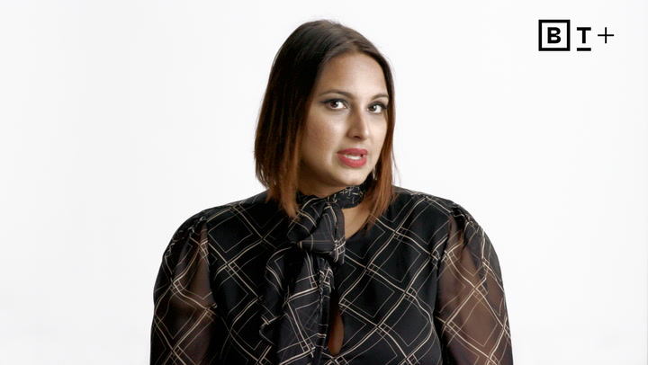 A person with straight brown hair and a patterned black blouse is speaking in front of a plain white background with a "BT+" logo in the top right corner.