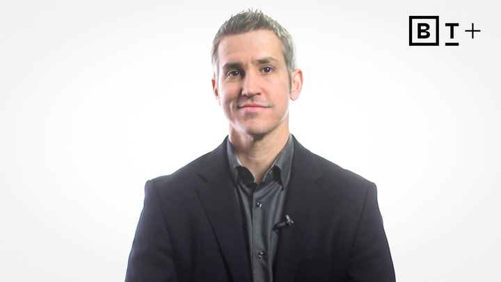 A man in a dark suit jacket and shirt sits facing the camera against a plain white background with a "B T +" logo in the upper right corner.