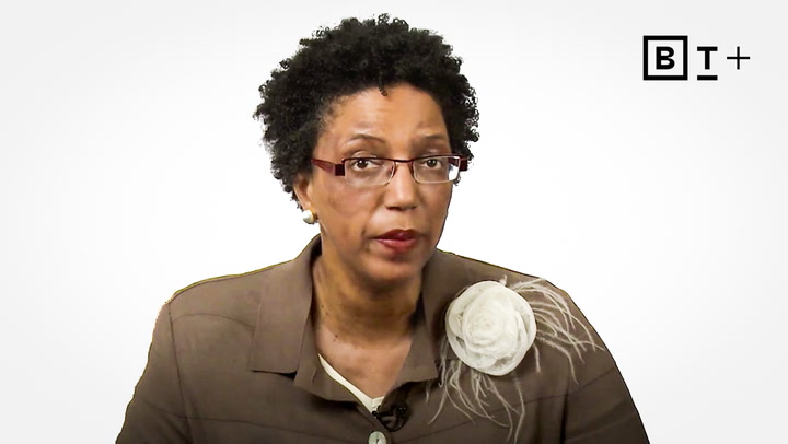 A woman with short curly hair and glasses wears a brown jacket with a white floral brooch, seated against a plain white background with a "B T +" logo in the top right corner.
