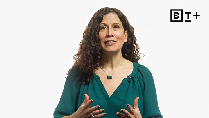 A woman with long curly hair speaks, gesturing with her hands. She wears a green blouse and a necklace. The letters "B T +" appear in the upper right corner on a plain white background.